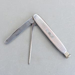 May include: A silver pocket knife with a blade and a tool for packing tobacco. The knife has a textured surface and a small hole at the end of the handle.