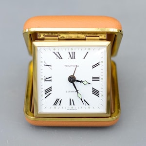 以下が含まれることがあります： 正方形の白い文字盤、黒いローマ数字、金色の針を備えたヴィンテージのトラベルクロック。 時計は、金色のフレームが付いたタン色の合皮ケースに収められています。 時計の文字盤には「TEMPORA」と「2 JEWELS」と書かれています。
