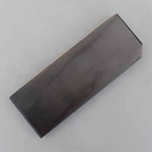 Puede incluir: Un objeto rectangular de madera marrón oscuro con un borde biselado. La veta de la madera es visible y la superficie parece lisa y pulida. El objeto está sobre un fondo gris neutro. La forma sugiere que podría ser un elemento decorativo o un componente de un objeto más grande.