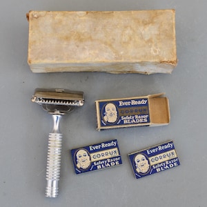 Może przedstawiać: Zabytkowa maszynka do golenia z srebrną rączką i główką, w zestawie z pudełkami z ostrzami. Pudełka z ostrzami są niebiesko-białe, z napisem "Ever-Ready CORR-U-X Safety Razor BLADE". Maszynka i ostrza leżą na szarej powierzchni.