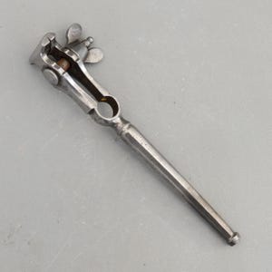 May include: A vintage metal hand vise tool featuring a long, slender handle, a circular opening in the center, and a clamping mechanism at the top controlled by a threaded wing nut. The tool has a weathered, dark grey metallic finish throughout.