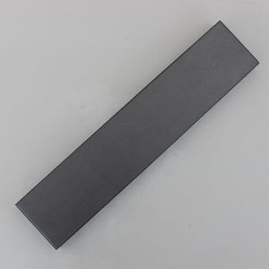 May include: A long, rectangular, dark gray box. The box is empty and has a matte finish. The box is angled diagonally across a light gray surface. The box is likely for packaging small items.
