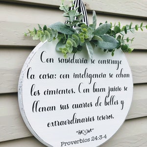 May include: White round wooden sign with black text that reads "Con sabiduría se construye la casa: con inteligencia se echan los cimientos. Con buen juicio se llenan sus cuartos de bellos y extraordinarios tesoros. Proverbios 24:3-4". The sign is decorated with green leaves and a black and white ribbon.