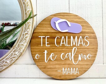 May include: A round wooden sign with the text "TE CALMAS o te calmo - MAMA" in white script. A purple flip-flop with a white strap is attached to the top. The sign is a decorative item.