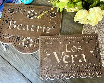 May include: Two wooden signs with cut-out designs and the names "Beatriz" and "Los Vera" written on them. The signs are brown and have a rustic look.
