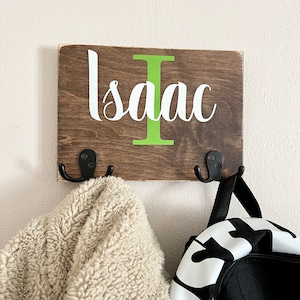 May include: Wooden coat rack with two hooks, painted brown with a green letter "I" and the name "Isaac" in white lettering. A beige fleece jacket and a black backpack with a white Nike logo are hanging on the hooks.