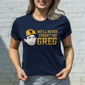 May include: Navy blue t-shirt with the text "WE'LL NEVER FORGET YOU GREG" in yellow. The shirt features an illustration of a person wearing a yellow and blue hat with the outline of Wisconsin. The shirt is a crew neck.