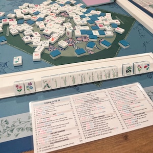 May include: A Mahjong game set featuring a blue and green patterned play mat, a white plastic tile rack holding numbered and floral tiles, a scattered pile of white and blue tiles, and a printed Mahjong On The Go 2020 score card with rules.