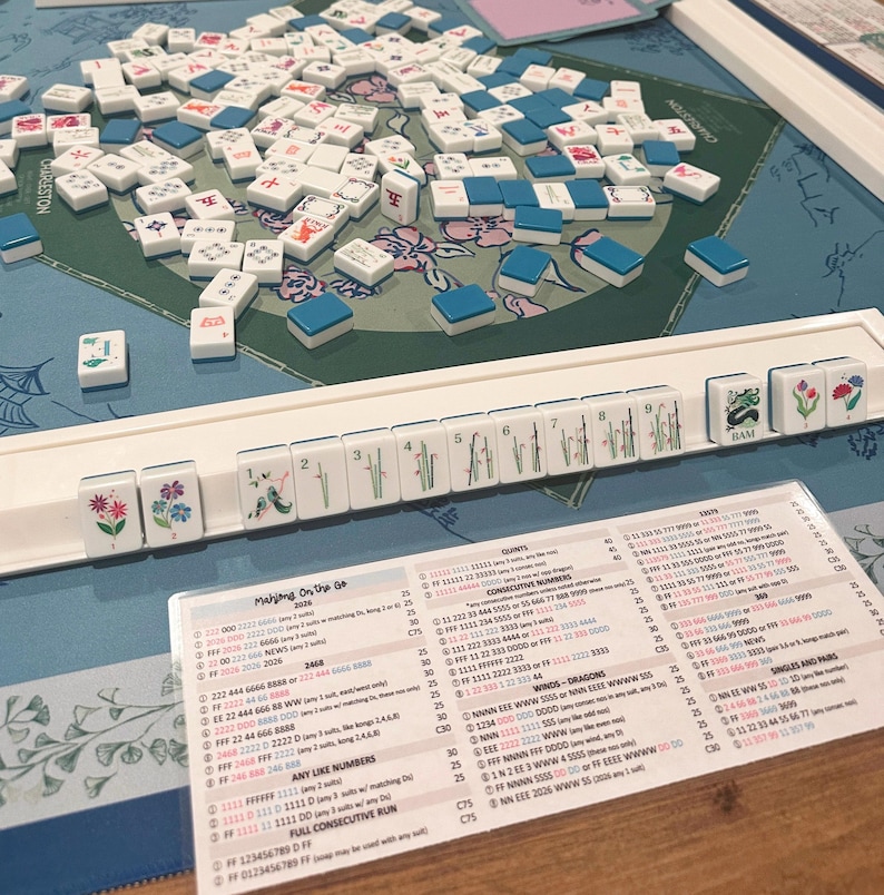 May include: A Mahjong game set featuring a blue and green patterned play mat, a white plastic tile rack holding numbered and floral tiles, a scattered pile of white and blue tiles, and a printed Mahjong On The Go 2020 score card with rules.