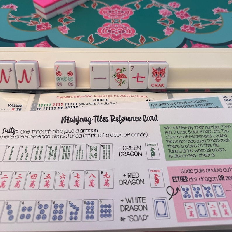 K&ouml;nnte beinhalten: Eine Mahjong-Fliesen-Referenzkarte mit gedruckten Abbildungen von Farben, Drachen und Spielregeln, platziert hinter einem wei&szlig;en Fliesenhalter, der mehrere wei&szlig;e Fliesen mit roten und gr&uuml;nen Symbolen enth&auml;lt, darunter ein Flamingo und ein Drachenzeichen.