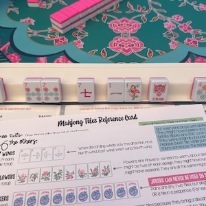 K&ouml;nnte beinhalten: Ein Mahjong-Spielset mit wei&szlig;en Spielsteinen mit rosa R&uuml;ckseite auf einer t&uuml;rkisfarbenen Blumenmatte. Ein wei&szlig;er Spielsteinhalter zeigt Steine mit roten, gr&uuml;nen und schwarzen Markierungen. Eine Mahjong-Referenzkarte mit Text und Illustrationen.