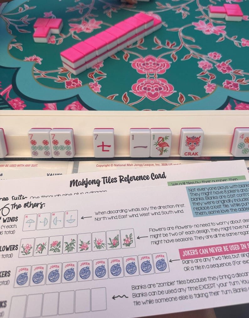 K&ouml;nnte beinhalten: Ein Mahjong-Spielset mit wei&szlig;en Spielsteinen mit rosa R&uuml;ckseite auf einer t&uuml;rkisfarbenen Blumenmatte. Ein wei&szlig;er Spielsteinhalter zeigt Steine mit roten, gr&uuml;nen und schwarzen Markierungen. Eine Mahjong-Referenzkarte mit Text und Illustrationen.