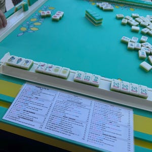 May include: A Mahjong set featuring white tiles with green backs and colorful illustrations, arranged on a teal playing surface with a lemon pattern. A printed score card with columns of text and numbers is placed in front of the tile racks.