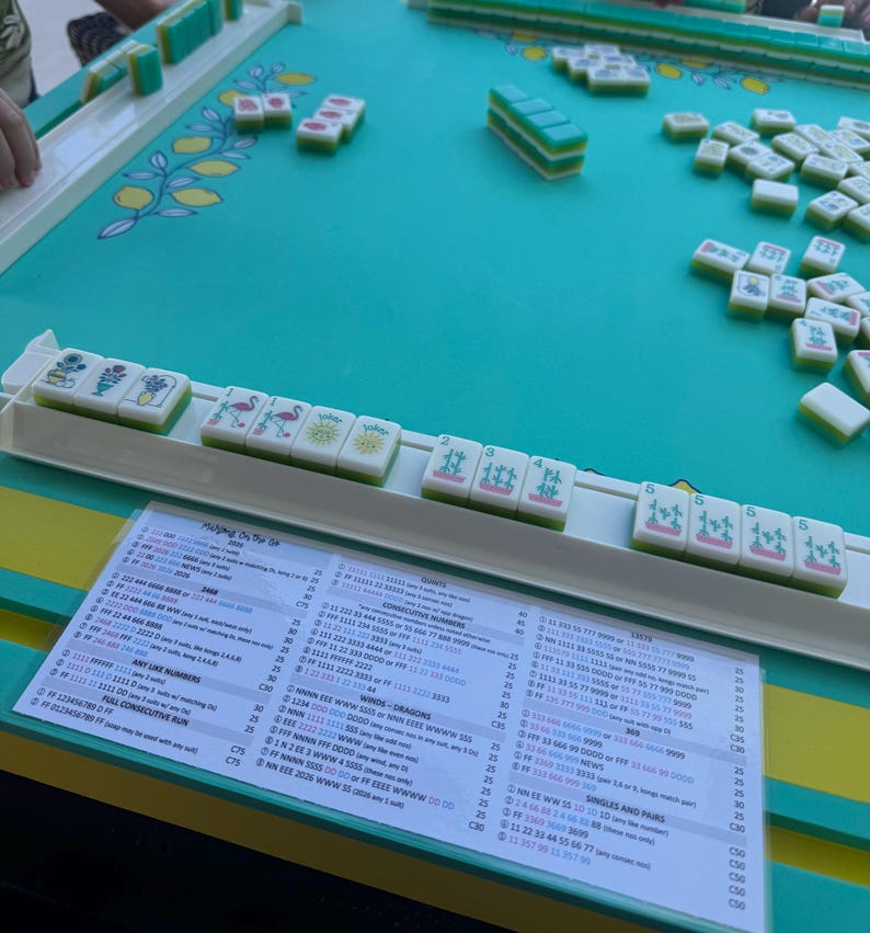 May include: A Mahjong set featuring white tiles with green backs and colorful illustrations, arranged on a teal playing surface with a lemon pattern. A printed score card with columns of text and numbers is placed in front of the tile racks.