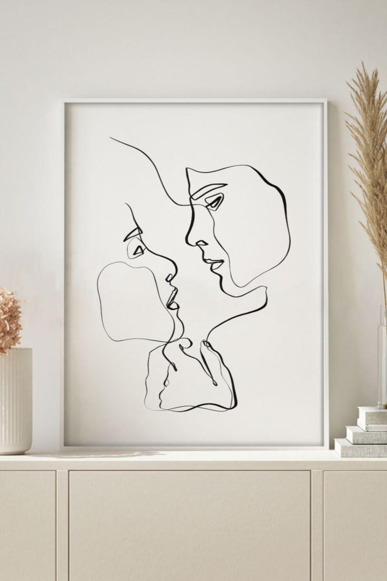 May include: Black and white line art print of two faces in profile, nearly touching. The minimalist artwork is framed in a white border, creating a modern aesthetic. The artwork is displayed on a white cabinet, with decorative elements in the background.