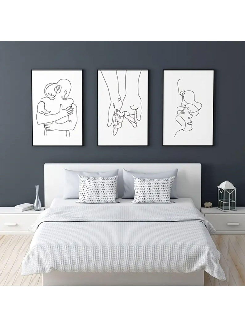 May include: Three framed black and white line art prints depicting human figures. The art pieces are displayed above a bed with white bedding and decorative pillows. The wall behind the art is a dark gray color.
