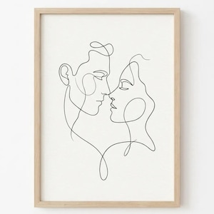 May include: A framed minimalist line art print featuring two faces in profile, almost touching. The black lines create the outlines of a man and a woman, set against a white background. The artwork is in a light-colored wooden frame.