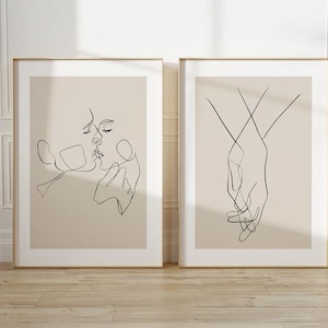 May include: Two framed art prints. The left print features a line drawing of two faces kissing. The right print shows two hands holding each other. Both prints have a beige background and are in gold frames.
