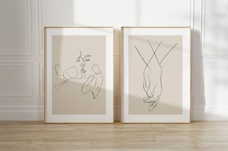 May include: Two framed art prints. The left print features a line drawing of two faces kissing. The right print shows two hands holding each other. Both prints have a beige background and are in gold frames.