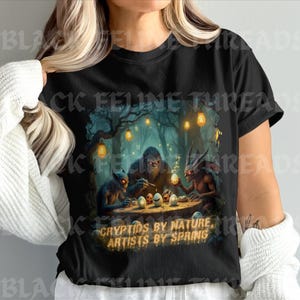 May include: Black t-shirt featuring a fantasy scene with cryptids around a table. The image includes a Bigfoot-like creature and other mythical beings, with the text "CRYPTIDS BY NATURE, ARTISTS BY SPRING".