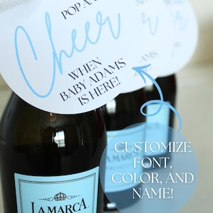 May include: Bottles of La Marca Prosecco sparkling wine with custom baby shower tags. The tags are white with blue text that reads "Pop and Cheer when Baby Adams is Here!" and are tied with white ribbon.
