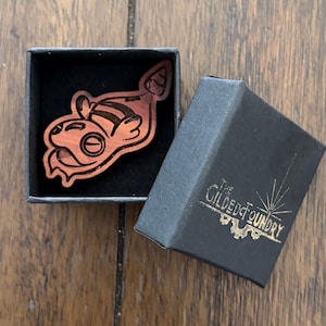 May include: A wooden pin in the shape of a cartoon character, resting in a black box with a black interior. The box is open, revealing the pin's details. The box has gold text that reads "The Gilded Foundry".