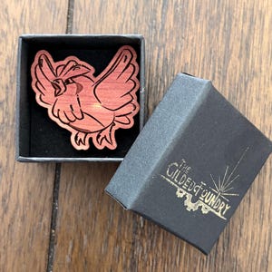 May include: A wooden pin in the shape of a bird with outstretched wings, resting in a black box. The pin is reddish-brown with etched details. The box is open, revealing a black interior. The box lid has gold text that reads "The Gilded Foundry".