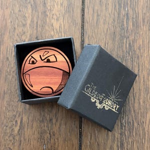Può includere: Una spilla rotonda in legno con un design a faccia arrabbiata, presentata in una scatola nera. La scatola è aperta, rivelando i dettagli della spilla. La scatola ha il testo "The Gilded Foundry" in lettere dorate.