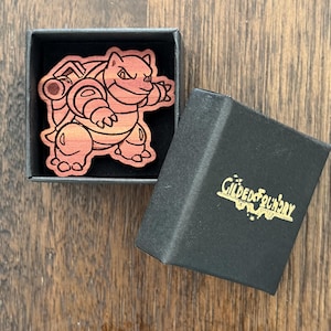 May include: A wooden pin shaped like the character Blastoise with a reddish-brown gradient finish, resting inside a small black square gift box. The lid of the box is detached and features the gold-colored text Gilded Foundry printed on the top.