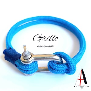 Op de afbeelding: Een handgemaakte blauwe nautische armband met een roestvrijstalen sluiting. De armband heeft het woord "Grillo" en het woord "handmade" in zwart schrift. De armband is gemaakt van een dik, geweven touw.