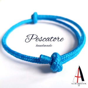 Puede incluir: Un brazalete de cuerda azul brillante con un nudo en cada extremo. El brazalete está hecho a mano y tiene el texto "Pescatore handmade".