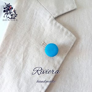 Può includere: Un piccolo bottone in tessuto turchese brillante con uno spillo d'argento, utilizzato come spilla da revers. Lo spillo è su uno sfondo in tessuto beige. Il testo "Riviera handmade" è sotto lo spillo.