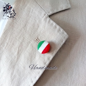 May include: A handmade Italian flag lapel pin featuring green, white and red colours. The pin is attached to a beige fabric background with the word "Handmade" written below it.