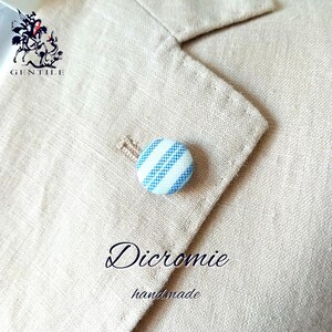 May include: A light beige blazer with a blue and white striped button. The button is round and has a textured appearance. The word "Dicromie" and "handmade" are written in cursive on the blazer.
