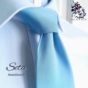 Può includere: Una cravatta azzurra annodata, indossata sopra una camicia bianca con colletto. L'immagine presenta il testo GENTILE con un logo di un cavaliere nell'angolo in alto a destra e le parole Seta handmade nell'angolo in basso a sinistra.