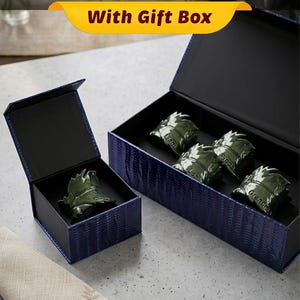 May include: A set of green leaf-shaped napkin rings inside a navy blue gift box. The box has a textured pattern and a black interior. The napkin rings are engraved with names. A folded beige napkin is in the foreground.