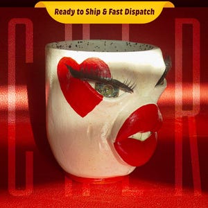 May include: White ceramic planter with a painted face, featuring a red heart, a green eye, and red lips. The planter is on a red surface.