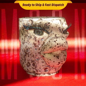 Puede incluir: Una taza de cerámica con un diseño de cara de luna. La taza tiene un esmalte moteado marrón y blanco y presenta una boca grande abierta con un labio blanco. Los ojos están pintados con pestañas negras e iris azules.