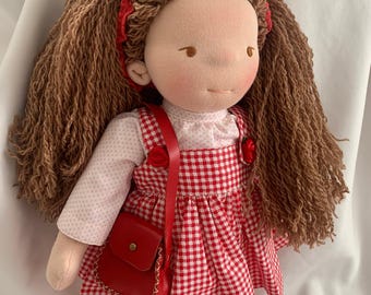 May include: A handmade cloth doll with long brown hair and a red headband. The doll wears a white blouse with a red and white checkered dress, a red purse, and red shoes. The doll's face is hand-painted with rosy cheeks.