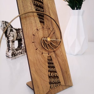Puede incluir: Un reloj de madera con un diseño único. Los números del uno al doce se muestran en espiral alrededor de la esfera del reloj. El reloj está hecho de madera marrón claro y tiene una aguja dorada.