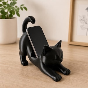 May include: A black cat-shaped phone stand featuring a feline in a stretching pose with its front paws extended forward and its tail curved upward. The device is held securely in a slot located on the back of the cat figure, providing a stable base.