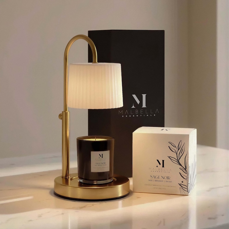 May include: A gold-toned candle warmer lamp with a white pleated shade and a black candle. The candle is in a black glass jar. A box with the text "SAGE NOIR" and "MALBELLA ESSENTIALS" is also present.