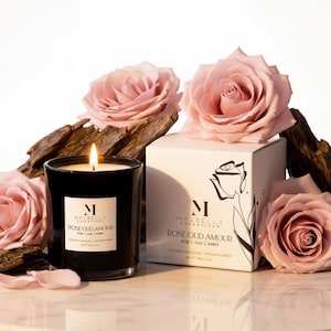 May include: A lit black candle in a glass jar with a white label that reads "Rose Oud Amour" is surrounded by pink roses and a white box with the same label. The candle is a coconut wax blend and weighs 14.8 oz.