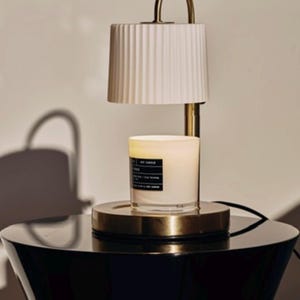 May include: A candle warmer with a white pleated lampshade and a gold-toned base. A lit candle sits on the base, with a black label. The warmer has a curved handle and is placed on a black table.