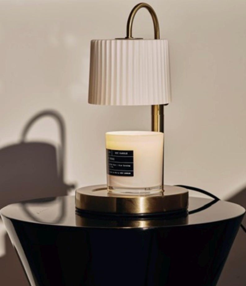 May include: A candle warmer with a white pleated lampshade and a gold-toned base. A lit candle sits on the base, with a black label. The warmer has a curved handle and is placed on a black table.