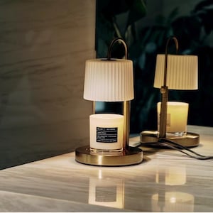 May include: Two candle warmers with a gold base and a cream-colored lampshade. The candle sits inside the warmer, and the lampshade is above it. The candle warmer has a black cord and plug. The background is dark with a marble surface.
