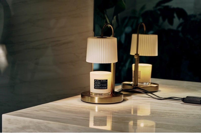 May include: Two candle warmers with a gold base and a cream-colored lampshade. The candle sits inside the warmer, and the lampshade is above it. The candle warmer has a black cord and plug. The background is dark with a marble surface.