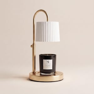 May include: A gold-toned metal candle warmer lamp featuring a curved arm and a white, vertically ribbed cylindrical shade. A black glass candle jar with a white label reading Malbella Essentials Rose Amour sits on the circular base.