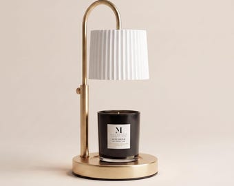 May include: A gold-toned metal candle warmer lamp featuring a curved arm and a white, vertically pleated cylindrical shade. Beneath the shade sits a black glass candle jar with a white label reading Malbella Rose Amour.