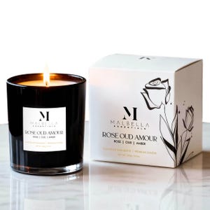May include: A lit black glass candle with a white label that reads "MALBELLA ESSENTIALS ROSE OUD AMOUR ROSE | OUD | AMBER". A white box with the same text and a rose illustration is next to the candle. The candle is 3.5 oz.